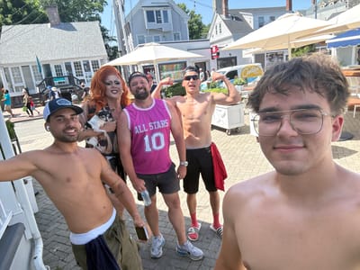 In a street scene in Provincetown, Massachusetts, USA, a group of five individuals is captured in a selfie on a sunny day. The backdrop consists of buildings with white siding and peaked roofs, typical of coastal New England architecture, along with outdoor seating areas shaded by large, off-white umbrellas.
In the foreground, closest to the camera and occupying the right half of the frame, is a young man with short, light brown hair and glasses. He is shirtless, and his shoulders and upper chest are visible.
To the left of this individual, another shirtless young man is positioned. He has short, dark hair and is wearing a black baseball cap. He is holding a small, white and black dog with a pink collar in his arms. Next to him, a person with vibrant orange hair and dramatic makeup, dressed in a black outfit with a floral print, is also visible.
Further to the left, a man with a beard and a pink tank top that reads "ALL STARS 10" is smiling and flexing his biceps. He is wearing dark shorts and athletic shoes. To his right, another man, also shirtless, is flexing his arms. He has a black baseball cap and sunglasses on, and is wearing black shorts with a red towel draped over his shoulder.
The ground is paved with brick-like stones. Other people are visible in the background, and a white van with "The Canteen" logo is parked behind the group. The atmosphere appears to be festive and casual, likely during an outdoor event or gathering.