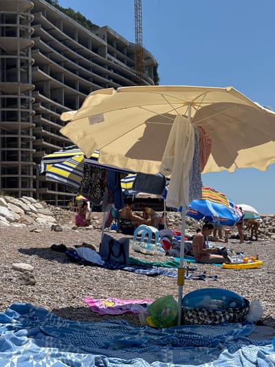 The image depicts a sunny day on a pebbled beach in Perazića Do, Montenegro, with an unfinished, multi-story concrete building under construction in the background. In the foreground, several beach umbrellas are set up, providing shade. One large, cream-colored umbrella is prominently featured, with clothing items hanging from its pole. Beach towels, bags, and inflatable toys are scattered on the ground around the umbrellas. People are visible in the background, some sitting under umbrellas, others potentially swimming or walking. The sea is not directly visible, but the presence of a beach implies its proximity. The overall atmosphere is one of a typical summer beach day, albeit with the unusual backdrop of a large construction project.