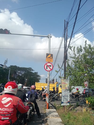 This media file captures a street scene in Surabaya, East Java, under a clear blue sky with scattered white clouds. The sun appears to be high, suggesting it's daytime. The image focuses on a busy intersection where numerous motorcycles and a few cars are present.

The primary subjects are people riding motorcycles, mostly wearing helmets. One prominent figure in the foreground wears a red jacket with "ACI Aku Cinta Indonesia" and "Food • Ride • Kurir" written on it, indicating a delivery service. Several other individuals are visible on their motorcycles, some with backpacks.

Objects in the scene include traffic lights, overhead wires, road signs, and a large yellow truck. One road sign clearly displays "AREA DIAWASI CCTV," indicating surveillance. Another sign shows a red circle with a diagonal line through a right-pointing arrow, meaning no right turn. A poster in the middle ground offers police assistance, providing a contact number for "POLDA JAWA TIMUR" and a "Call Center 110." Another poster advertises "Komunal P2P" for business capital, with contact information.

The setting is a typical urban intersection with roads, sidewalks, and vegetation on the side. The presence of traffic, signs, and a delivery service suggests an active and functional urban environment. The overall mood is that of a normal, bustling day in a city.