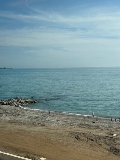 The image depicts a panoramic view of a coastline in Kholodnyi Rodnik, Russia, under a clear, hazy blue sky with scattered white clouds. The vast expanse of the turquoise-blue sea occupies the mid-ground, showing gentle ripples on its surface. On the distant horizon to the left, two large cargo ships are visible. The foreground features a beach composed of both gravelly sections near the water and a broader expanse of disturbed, light brown earth/sand closer to the viewer. Prominent tire tracks traverse this sandy area. Along the left edge of the beach, a substantial formation of large grey rocks extends into the sea, forming a partial breakwater.
Several individuals are present on the beach near the water's edge. To the left, near the rocky area, one person is partially submerged in the water. Further to the right, one individual in a light top is bending over near the water, while another stands facing the sea. Closer to the right side of the frame, three people are seen reclining or sitting on the pebbly shore. Two dark, circular objects, possibly tires, are positioned on the sand between the lounging individuals and the disturbed foreground. A low, white concrete barrier is partially visible in the bottom-left corner of the frame. The overall scene suggests a warm, sunny day with minimal human activity on the beach.