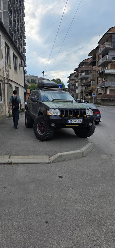 A black Jeep Grand Cherokee is parked on a street in Gori, Georgia. The vehicle has been heavily modified with off-road tires, a roof-mounted cargo box, and an LED light bar on the hood. The license plate reads "XD 368-DD". A person wearing a backpack and dark pants is walking away from the Jeep. In the background, there are apartment buildings with balconies and a cloudy sky. The time of day appears to be late afternoon, given the soft lighting. The scene is urban, with a mix of older and newer architecture visible.