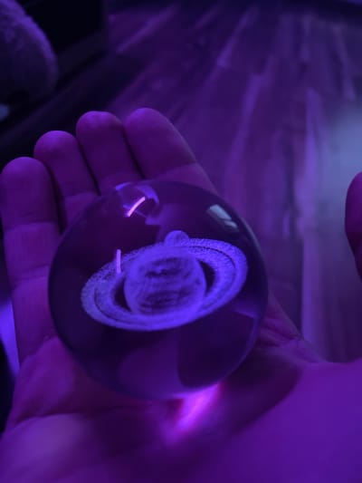 A hand is holding a glass orb containing a miniature Saturn. The orb is lit from below with purple light, casting a glow on the hand and the surrounding area. The background is dark and blurred. The scene appears to be indoors, with the purple lighting suggesting it could be a room with mood lighting or a specific display. There is no indication of the location being in Gliwice, Poland.