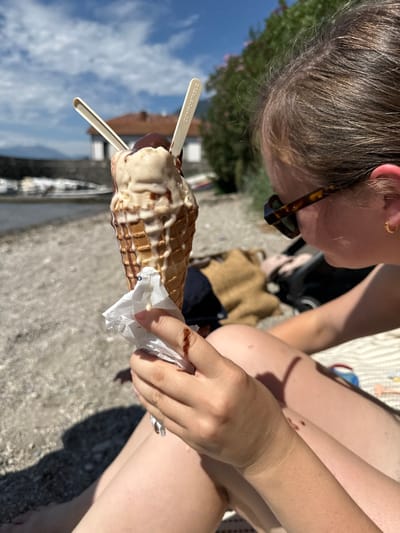 The image depicts a person holding a melting ice cream cone on a sunny day at a gravelly shoreline in Abbadia Lariana, Italy, overlooking Lake Como. The individual, seen from the right side, has brown hair and wears tortoiseshell-patterned sunglasses and a small gold hoop earring on the visible ear. Their right hand holds a waffle cone containing two scoops of ice cream—one light-colored and one dark-colored, likely chocolate—both actively melting and dripping down the cone's sides. Two small, light-colored spoons, explicitly marked "COMPOSTABLE," are inserted into the ice cream. A crumpled white paper napkin is wrapped around the base of the cone, soiled by the melted drips. The person's bare legs are visible, resting on a light-colored patterned textile. In the background, the calm waters of Lake Como feature several moored boats. Distant elements include a stone bridge, buildings with red-tiled roofs, and a mountainous landscape under a clear blue sky with scattered white clouds.