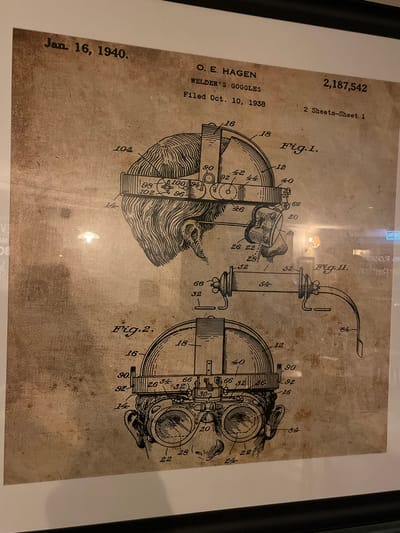 The image displays a framed patent document, presented on an aged, sepia-toned background with black line drawings and text. The document is titled "WELDER'S GOGGLES," issued to "O. E. HAGEN" on "Jan. 16, 1940," with a filing date of "Oct. 10, 1938," and patent number "2,187,542." It is identified as "2 Sheets-Sheet 1." The document features three distinct illustrations: "Fig. 1" depicts a side view of a head wearing adjustable goggles, showcasing the head strap, ear section, and the goggle mechanism. "Fig. 2" provides a front view of the goggles positioned on a head, detailing the dual lenses and forehead strap. "Fig. 11" illustrates a detached cylindrical component with connecting arms, possibly an accessory for the goggles. The document is enclosed within a dark frame with a white inner mat. Reflections from an interior environment, including a prominent circular light, are visible on the protective glass, slightly obscuring portions of the print. The image does not contain any discernible features or information that specify its exact location within the city of Bath, United Kingdom.