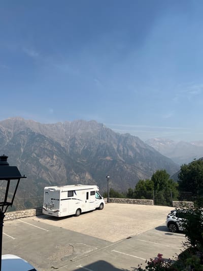 The image depicts a panoramic view of a mountainous landscape under a clear blue sky with sparse white clouds. In the foreground, a paved parking lot is visible. A white motorhome is parked in the lot, facing away from the camera, with a bicycle rack attached to its rear. To the right, a white hatchback car is parked, partially obscured by foliage. A black lamp post stands to the left of the motorhome. A stone wall borders the parking lot, beyond which are dense green trees and a metal railing. The background is dominated by a vast mountain range with rugged, rocky peaks, some of which are tinged with snow. The location is identified as the city of Benasque, Spain.