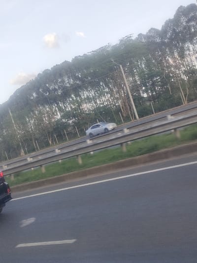 The image depicts a multi-lane asphalt highway within Juja ward, Kenya, under a light blue sky with scattered white clouds. A dense line of tall, slender trees, consistent with eucalyptus species, borders the highway on the right side. A median strip, featuring green grass and a metal guardrail barrier, separates opposing directions of traffic. In the foreground, the rear section of a dark-colored vehicle is visible on the left lane, suggesting forward movement. On the opposing carriageway, separated by the median, a silver sedan, possibly a Mercedes-Benz model, is in motion, heading away from the observer. White lane markings are visible on the asphalt. No individuals are discernible in the frame. The lighting indicates daytime conditions, possibly early morning or late afternoon.