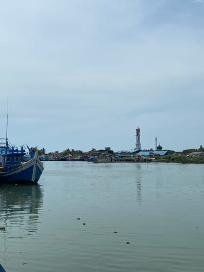 A sideways medium shot shows a view of Tanjongkapai, Indonesia, across a body of water, from the perspective of a boat.
The foreground features the bow of a blue and white boat with a visible antenna. The water is calm, with slight ripples and reflections of the cityscape.
Across the water, the cityscape includes buildings, houses, and greenery along the shoreline. Prominent features include a white mosque with a tall minaret topped with a golden dome, and another minaret in the distance. The sky is overcast.
