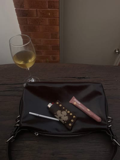 The image displays a still-life arrangement on a dark, weathered wooden table, located in Thirroul, Australia. In the upper left quadrant, a stemmed clear glass contains approximately half a serving of pale yellow liquid, consistent with white wine, and exhibits condensation on its exterior. Centered in the lower half of the frame, a dark brown, possibly patent leather, handbag or clutch lies flat. Resting on the handbag, from left to right, are a white filter cigarette with a partially visible "KENT" brand logo near the filter, a lighter housed in a dark brown, studded sleeve featuring a lighter-colored cross-stitch pattern, and a tube of "rhode Peptide Lip Tint" in a light pink hue. The background features a segment of a reddish-brown brick wall on the left, and a plain, light-colored door with a silver doorknob or handle on the right. The overall setting suggests an outdoor or semi-outdoor environment such as a patio or balcony. No individuals are present in the scene.