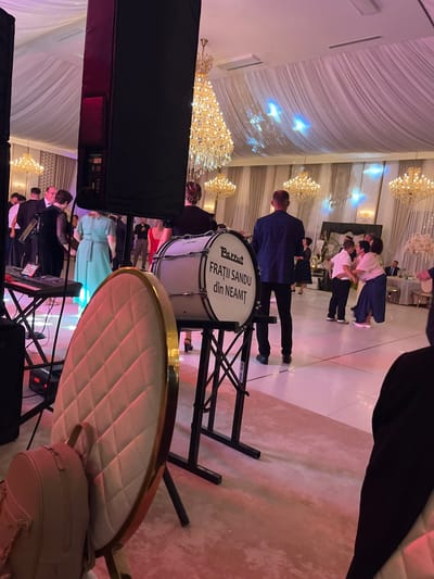 The image depicts a formal indoor event, likely a wedding reception or a large party, taking place in Huși, Romania. A band is set up on a dance floor. In the foreground, a large bass drum with the inscription "PARROT FRATII SANDU din NEAMȚ" is prominently displayed on a stand. To the left of the drum, a keyboard is set up on a foldable stand, with a tablet or screen attached. Behind the band setup, a quilted, gold-trimmed chair is partially visible, with a light-colored backpack resting on it.
In the midground, several individuals are present. A man in a dark suit is standing behind the bass drum, facing away from the camera. To his right, several couples and groups of people are interacting on the dance floor. One couple is embracing, while others are standing and conversing.
The background features a banquet hall with elegant decor. Draped white and grey fabric adorns the ceiling, with multiple large crystal chandeliers illuminating the space. Soft, colored lighting, predominantly pink, casts a warm glow across the scene. The walls are covered in textured panels and decorative curtains. Tables with chairs are visible in the distance, suggesting a dining area or lounge space.
The overall atmosphere appears festive and celebratory, with elements of a live musical performance and social gathering.