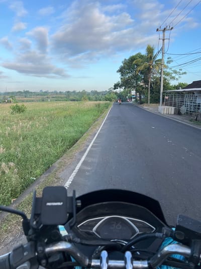 The image is a first-person perspective from a motorcycle on a paved road in Abiansemal, Indonesia. The road is lined with green fields and vegetation on the left and buildings and utility poles on the right. The sky is partly cloudy with a clear blue expanse above. In the foreground, the handlebars and dashboard of the motorcycle are visible, with a phone mount and a digital speedometer displaying "36". In the middle ground, there are a few vehicles on the road, including a scooter and a red car. The background features more greenery, palm trees, and scattered buildings, suggesting a rural or semi-rural setting. The overall scene depicts a serene journey through the Indonesian countryside.