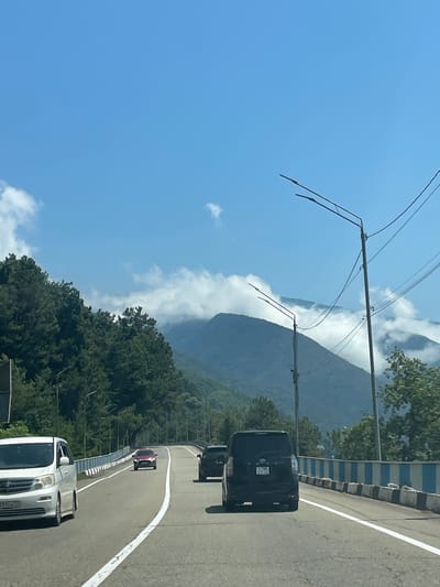 In Tsandrypsh, Abkhazia, a road winds through a mountainous landscape under a clear blue sky dotted with clouds. Several vehicles are on the road, including a white minivan on the left, a red car further ahead, and two dark minivans. The road is bordered by a guardrail with blue and white panels. Lush green trees line the hillsides, and the peaks of the mountains are partially obscured by clouds. Streetlights and power lines are visible along the road. The scene depicts a scenic drive through a region known for its natural beauty.