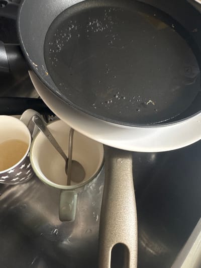 The image displays an overhead view within a metallic kitchen sink. The primary subject is an inverted frying pan with a dark, non-stick interior showing minor wear near the rim and containing a shallow layer of clear liquid with numerous small bubbles and sparse particulate debris. The pan's exterior is light-colored, and its speckled metallic grey handle, featuring a hanging hole, extends downwards out of the frame.

Adjacent to and beneath the pan are two ceramic mugs. To the left, a white mug with a dark, star-like pattern on its exterior holds a light amber liquid. Centrally positioned beneath the pan's rim is a light green-grey mug with a white interior, containing two metallic utensils: one spoon and one slender, elongated implement, possibly a stirring rod or small ladle. The sink basin is wet, consistent with recent use or washing.

There are no people visible. The scene depicts kitchenware in a sink, suggesting cleaning or preparation activities. The image provides no discernible visual information to establish its location in the city of Visp, Switzerland.