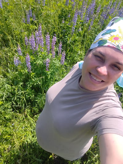 A woman is taking a selfie in a field of lupines. She is wearing a grey t-shirt and a headscarf with a floral pattern. The field is green with tall purple lupine flowers. The background shows more lupines and greenery. The woman is smiling and looking at the camera. The lighting suggests it is a sunny day.
