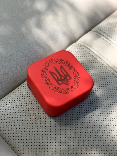A close-up, high-angle shot shows a red, square-shaped case with a black emblem of a trident, surrounded by a wreath of leaves. The case sits on a perforated light gray surface, likely a car seat, with visible stitching and a textured pattern. Sunlight casts shadows on the surface. The image does not provide any information about the city of Southlake, United States of America, or the surrounding environment beyond the immediate object and its resting surface.