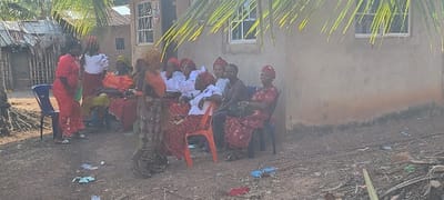 The image depicts an outdoor gathering of approximately 12 individuals, primarily adult females, situated in Alayi, Nigeria. The scene is illuminated by bright daylight, casting distinct shadows.
The ground is unpaved, consisting of dirt with scattered debris, including plastic items and wrappers. Overhead, palm fronds partially obscure the sky.
On the left side of the frame, a woman in a red lace dress stands, holding a light green object. To her immediate right, another woman stands wearing a white blouse with ruffled sleeves and a red patterned skirt, adorned with a matching head wrap. An empty blue plastic chair is positioned to the left of the woman in red. Behind these two, a woman in an orange top and red patterned skirt is seated.
Towards the center and right, a group of approximately seven women are seated on various plastic chairs, including orange and black models. Several individuals in this group wear coordinated outfits consisting of white blouses with ruffled sleeves, red patterned skirts or wrappers, and matching red head wraps. One seated woman wears a dark top, dark lower garment, and a dark head wrap, holding a dark bag. Another woman, seated on the far right, wears a red patterned dress and a red head wrap, also holding a dark bag. A woman in a patterned brown and red dress stands in the foreground, facing left, partially obscuring some seated individuals.
The background features two primary structures. On the right, a single-story building with a light brown, weathered exterior and two white-framed windows is visible; its lower wall shows dark staining. To the left, a more rudimentary structure with a corrugated metal roof and walls constructed from woven materials or slats is present, with another building behind it. Assorted debris, including thin metallic rods, is scattered on the ground near the right-side building.