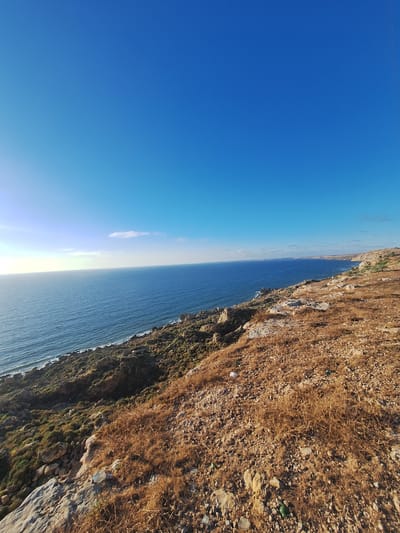 The image depicts a dramatic coastal landscape, likely near Ahrara, Morocco. The scene is split between a vast, clear blue sky and the deep blue ocean, which stretches to the horizon. The sun is low in the sky, casting a bright glare near the horizon line and suggesting it is either early morning or late afternoon. The weather appears to be clear and sunny.

To the right, a rugged, rocky cliff face dominates the foreground. The cliff is covered in dry, brown vegetation and scattered stones, giving it a parched appearance. The terrain is steep and uneven, with some areas showing patches of green growth, indicating a mix of arid and somewhat vegetated sections. A small, light-colored object, possibly a bottle or a piece of plastic debris, rests on the rocky surface of the cliff.

The ocean is calm with gentle waves lapping at the base of the cliff. The sunlight reflects off the water, creating a shimmering effect. The sky is almost entirely clear, with only a faint wisp of a cloud visible in the distance, high above the ocean. The orientation of the photo, with the sky on the left and the cliff on the right, is unusual and suggests the photo may have been taken with a handheld device and rotated. There is no visible text in the image.