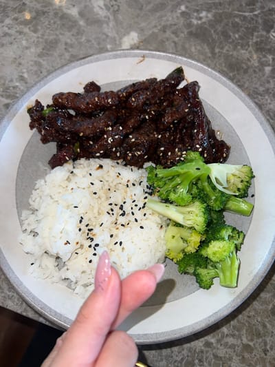 A close-up, overhead shot showcases a plate of food on a marble surface. The plate contains a portion of white rice sprinkled with black and white sesame seeds, a serving of vibrant green broccoli florets, and thinly sliced beef coated in a dark, glossy sauce, also topped with sesame seeds. A person's hand, with manicured fingernails, is visible in the foreground, making a heart shape with their thumb and index finger. The lighting is bright, highlighting the textures and colors of the meal. There are no discernible landmarks or context clues to determine a specific location in Abu Dhabi, United Arab Emirates.
