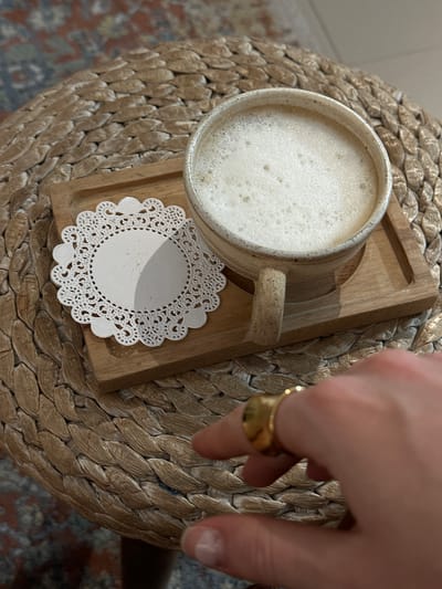 A high-angle shot displays a mug filled with a light-colored, frothy beverage set on a small wooden tray. A delicate white doily lies between the mug and the tray. The mug rests on a round, woven stool-like surface. A hand with a gold ring is partially visible in the upper right, appearing to reach towards the mug. The location is assumed to be in Kerobokan Kelod, Indonesia.