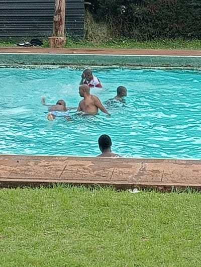 The image depicts an outdoor swimming pool scene at Riara Ridge, Kenya, where several individuals are enjoying the water during the daytime. The main subjects include an adult bald man with a bare torso, partially submerged in the clear, turquoise water, appearing to be among several children.
To the man's left, a child with dark hair is seen with an arm raised, seemingly holding a yellow object, possibly a ball or float. Another child, wearing a distinctive red and white striped top, is further back in the pool. A third child's head is visible to the adult's right. Additionally, a fourth individual is partially visible from the back in the lower foreground of the pool, with only their head and shoulders above the water line.
The pool itself features a light brown, textured coping around its edge. Surrounding the pool are elements of a natural outdoor setting: a strip of vibrant green grass occupies the foreground, and more lush green foliage and trees are visible in the background. A dark, corrugated metal fence or wall runs across the far background, behind some tree stumps.
The activity taking place is recreational swimming or playing in the pool. The scene suggests a relaxed and enjoyable event. The lighting indicates it is daytime, likely under overcast or soft sunlight, as no strong shadows are cast. There is no visible text in the image.