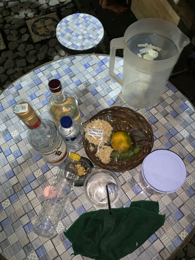 This image depicts an outdoor setting, possibly a casual dining area, with a table covered in a mosaic of blue, gray, and white tiles. The table is set with various items, suggesting a gathering or meal.

Prominently featured are several bottles, including one labeled "Captain Morgan Spiced Gold," another clear bottle with a red-brown cap, and a smaller bottle of water with a blue cap. A plastic jug filled with a clear liquid, possibly water or a beverage, sits on the table. A woven basket contains snacks, an orange, and a small packet of dark material, possibly a condiment or sweet. A clear plastic cup with a black straw rests beside the basket, and a large clear plastic water bottle, adorned with a pink flower-shaped object, lies on its side. A white lidded container and a dark green cloth are also present on the table.

The background shows a mix of rough gravel and what appear to be decorative mosaic tiles, suggesting an outdoor patio or garden area. In the upper left corner, a circular object with a similar tile pattern is visible, possibly a stool or another table. The lighting indicates it might be evening or night.

While the image does not explicitly contain identifying markers of Banjar, Indonesia, the presence of specific beverage brands and the overall ambiance are consistent with casual outdoor settings found in many Indonesian cities. However, without further contextual information, a precise location within Banjar cannot be definitively established. The image focuses on the arrangement of drinks and snacks, implying a moment of relaxation or social interaction.
