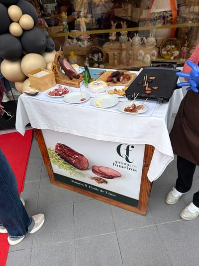 An outdoor food sampling station is positioned on a grey tiled pavement in front of a retail storefront in Felgueiras, Portugal. A rectangular table, covered with a white tablecloth, holds various food items. On the left, a whole cured ham (likely *presunto*) is mounted on a wooden stand, with thinly sliced pieces presented on a small white plate below it. Adjacent is a wooden box containing white napkins. In the center, several rounds of cheese, various types of cured sausages (some whole, some sliced), and a plate of bread or crackers are displayed. A small green bottle is visible behind these items. On the right, a black electric griddle is cooking small pieces of sausage, some with toothpicks inserted, and other cooked pieces are on small white plates.
Behind the table, the storefront window displays numerous decorative items, including Easter bunny figurines, teapots, and ceramic eggs, suggesting a gift or gourmet shop. A large arrangement of black and beige balloons is partially visible on the left, bordering the storefront. Beneath the table, a framed advertising sign depicts various cured meats with herbs, bearing the text "Quinta dos Fumeiros" and "Poiares Ponte de Lima."
The lower body of two individuals is visible. On the left, a person wears blue jeans and light-colored sneakers, holding dark sunglasses. A red floor mat is partially visible under their feet. On the right, a person wears a brown apron over dark trousers and white/gold sneakers; their right hand, clad in a blue disposable glove, is positioned near the electric griddle.