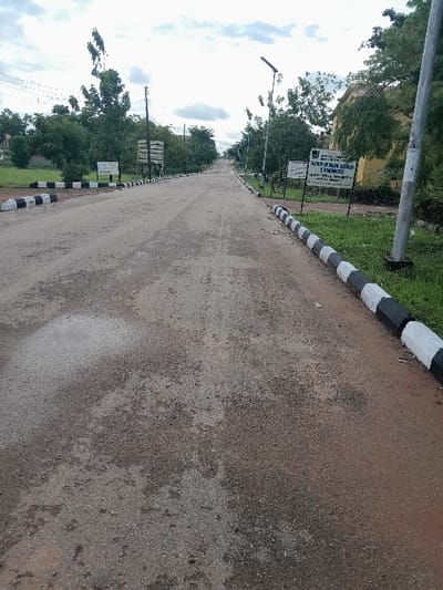 The image displays an asphalt road at the permanent site of Ebonyi State University in Ezzamgbo, Ebonyi State, Nigeria. The road, exhibiting signs of wear and minor surface irregularities with some damp patches, stretches linearly into the distance under an overcast sky. Both sides of the road are delineated by low concrete curbs painted with alternating black and white stripes. Adjacent to the curbs are strips of green grass and numerous trees. On the right side, a white signboard clearly states "EBONYI STATE UNIVERSITY, ABAKALIKI" and "PERMANENT SITE, ABAKALIKI". A solar-powered streetlight post equipped with a solar panel is also visible on the right, along with a partial view of a light-colored building beyond the trees. Utility poles with overhead wires are present along both sides of the road. No individuals are visible in the scene.