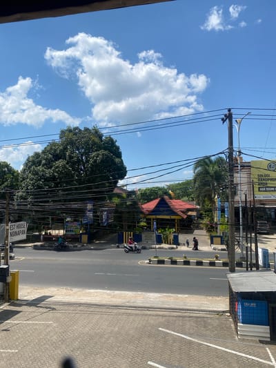The image shows a street scene in Keraton, Indonesia, on a sunny day with scattered white clouds. The foreground features a paved walkway with brick-like patterns and white parking lines. To the right, a blue structure with the text "GALA JAYA" is visible, partially enclosed by a black metal fence. The main street occupies the middle ground, with a few motorcycles on the road. On the left side of the street, a sign reads "EST HOUSE NGINAPAN". Across the street, a large, lush green tree dominates the landscape, with its branches obscuring a building with a red roof. Several utility poles and a dense network of overhead wires are prominent throughout the scene. In the background, there are more trees and buildings, including a yellow building with the text "SOLUSI HANDPHONE ANDA ADA DISINI". A few people are walking on the sidewalk. The overall environment suggests a commercial or residential area in a tropical setting.