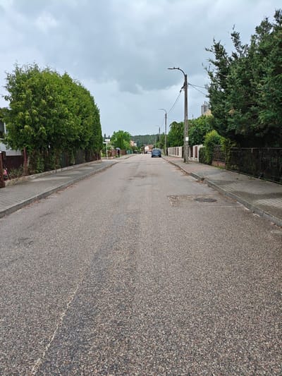 The image shows a street in Mława, Poland, on a cloudy day. The street is paved with asphalt and has concrete sidewalks on both sides. There are houses and fences lining the street, with mature trees and bushes providing greenery. A few streetlights are visible along the road. A blue car is parked on the left side of the street in the distance. A person holding a black umbrella is walking on the sidewalk on the right side of the street. The sky is overcast with grey clouds.