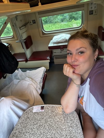 A young woman with blonde hair pulled back and wearing a purple and white t-shirt rests her chin on her hand, smiling at the camera. She is sitting in a train compartment, with a window to her left showing a green, tree-filled landscape. In the foreground, a speckled gray table is visible, and to her right, a white duvet is spread out. The train compartment appears to be a sleeping berth, with burgundy seats and upper bunks visible. The overall scene suggests a comfortable journey on a train. The location of Баграш-Бигра, Russia, is not evident from the image itself, as there are no identifying landmarks or signs within the frame that would indicate a specific city.