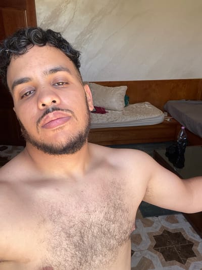 A selfie-style portrait shows a young man of medium skin tone, rotated 90 degrees counter-clockwise. He has curly dark hair, dark eyebrows, brown eyes, and a full beard and mustache. His expression is neutral as he looks directly at the camera. He is shirtless, revealing a significant amount of dark body hair on his chest.
The background is a mix of indoor elements. To his left (or above his head in the original orientation), there is a light-colored, textured wall. To his right (below his body in the original orientation), a patterned floor is visible, featuring geometric star-like designs in shades of brown, beige, and white tiles.
Further into the background, a wooden door or cabinet is present. Near this wooden surface, there are cream or white pillows and what appears to be the edge of a mattress or bed. Above his right arm, a clear plastic bottle is seen, next to a crumpled black plastic bag. The lighting suggests it is daytime. The setting is an indoor room, likely a residence in Baraki, Algeria. There is no visible text in the image.