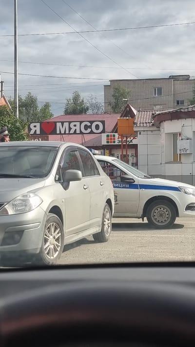 In Megion, Russia, a silver sedan is parked next to a police vehicle. The police car has a blue stripe along its side and the word "ПОЛИЦИЯ" (POLITSIYA) written on it. Behind the vehicles, a building with a red roof has a sign that says "Я ♥ МЯСО" (YA LYUBLYU MYASO, meaning "I Love Meat"). There is a traffic light visible in the background. The weather appears to be overcast.