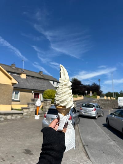 A hand in a black sweater and with dark nail polish holds a soft-serve ice cream cone in a waffle cone. The ice cream is white and has a swirled texture. The hand is holding a white paper napkin around the base of the cone. The scene is set outdoors on a sunny day, with a bright blue sky and some wispy white clouds. In the background, there are buildings with yellow and grey facades, a stone wall, and several cars parked along a street. Trees and a street light are also visible. The overall mood appears to be cheerful and relaxed, consistent with enjoying an ice cream on a nice day. The image is oriented vertically, with the sky occupying the top portion and the ground and buildings in the lower portion. This appears to be a street scene, possibly in a town or village. Based on the provided location context, it is likely Westport, Ireland. There is no visible text in the image.
