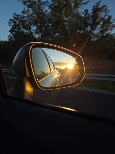 The image captures a sunset reflected in a car's side-view mirror while driving. The reflection shows a road stretching into the distance with the sun low on the horizon, casting a warm glow. Trees line the side of the road. The car's side mirror housing is visible in the foreground. The image is taken from inside the car, looking out. The location is the outskirts of Rajana, Bulgaria.
