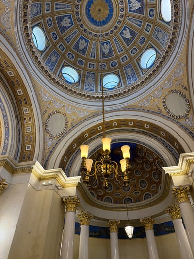 The image shows the interior of a grand dome, likely in a public building such as a museum, government building, or religious site, located in посёлок дачного хозяйства «Архангельское», Russia. The dome is elaborately decorated with intricate patterns and motifs in gold and blue. Oval windows are visible in the upper part of the dome, allowing natural light to filter in. A multi-tiered chandelier hangs from the center of the dome, illuminating the space with warm light from its globes. The lower parts of the image reveal white columns with ornate gold capitals, supporting the structure. The overall impression is one of opulence and historical significance.