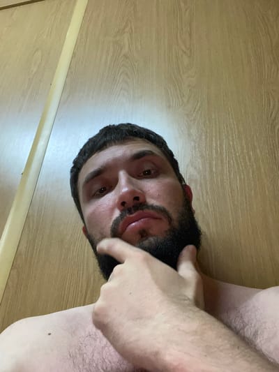 A close-up, low-angle, indoor selfie shows a man with dark hair and a beard resting his chin on his hand. He is positioned against a wooden-grained wall with a vertical light-colored strip on the left side. The man is shirtless, and his skin appears to have light hair. The lighting is warm, creating shadows on his face and suggesting an indoor setting. No specific location in a city or country can be determined from the image.