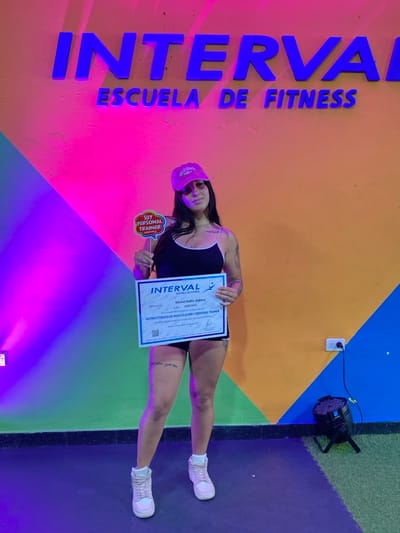A female subject stands centrally in an indoor setting, identified as Interval Escuela de Fitness in Quilmes, Buenos Aires, Argentina. She wears a black tank top, dark shorts, light-colored high-top sneakers, and a pink baseball cap with "California" embroidered in white. Tattoos are visible on her left arm and both upper thighs. She holds two items: a red speech bubble sign on a stick in her right hand (viewer's left) that reads "SOY PERSONAL TRAINER," and a white certificate in her left hand (viewer's right). The certificate, issued by Interval Escuela de Fitness, is for "INSTRUCTORADO DE MUSCULACIÓN Y PERSONAL TRAINER" for "Michel Nahir Aldana." The background wall is painted in a vibrant, multi-color gradient from left to right: magenta/purple, orange, and light blue/teal, with large blue raised letters spelling "INTERVAL" and "ESCUELA DE FITNESS." A light fixture illuminates the left side of the wall. On the lower right, a black stage light rests on the floor, which consists of dark gray and green sections. Two white power outlets are visible on the orange wall section. The subject is looking directly at the camera with a slight smile.
