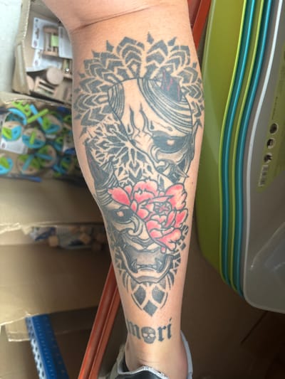 A leg tattoo, primarily black and gray with accents of red, is visible on a person's calf. The tattoo depicts a Japanese demon mask (Oni) with a red flower, possibly a peony, superimposed over it. Below the mask, the word "mori" is tattooed in black, with a small skull preceding it. The leg is positioned in a somewhat cluttered environment. To the left of the leg, partially visible, are cardboard boxes containing what appear to be children's building blocks or toys. To the right, stacks of brightly colored plastic containers, predominantly green and teal, are present. The background suggests an indoor setting, possibly a storage area or a workshop, with a floor that appears to be made of wood or laminate. The lighting suggests it is daytime, possibly with natural light filtering in. The exact location in the city of Kazichene, Bulgaria, cannot be determined from the visual information provided in the image.