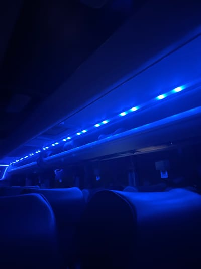 The image shows the dimly lit interior of what appears to be a public transport vehicle, such as a bus or train, viewed from a low angle looking upwards and along the aisle. The predominant feature is a long strip of bright blue LED lights running along an overhead panel or ceiling structure on the left side of the frame, extending into the distance. These lights are the primary source of illumination, casting a deep blue hue over the entire scene and creating a dark, atmospheric ambiance.
On the right side of the image, several dark, upholstered seats are partially visible, angled slightly away from the viewer. The subtle reflection of the blue light highlights their rounded backs and contours against the surrounding shadows. Above and beyond the seats, some indistinct fixtures, potentially luggage racks or ventilation grilles, are barely discernible in the darkness. No people are visible, and no specific activity is taking place.
The scene suggests travel during night-time or in very low light conditions, given that the LED lighting is the sole source of illumination. The cool blue lighting creates a serene or futuristic mood. There is no visible text within the image. The vehicle is consistent with modern coaches or trains found in regions such as Woking, United Kingdom.