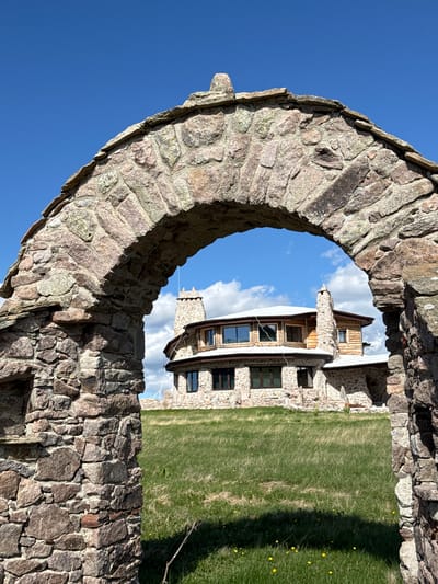 The image captures a grand stone archway in the foreground, framing a view of a large, multi-level stone and wood house in the background. The house features multiple chimneys and numerous windows. The surrounding environment is a grassy field under a bright blue sky dotted with white clouds. The image does not depict the city of Gela, Bulgaria.