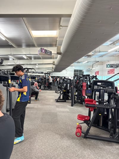 A person in a blue and yellow jersey performs a push-up in a gym. Exercise machines line the gray carpeted floor. A long cylindrical vent runs the length of the room. Signs for "Functional Area" and "Chest & Arms" are visible. Other people are using the equipment in the background.