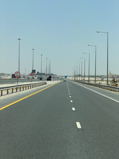 A wide, multi-lane highway or expressway stretches into the distance under a clear, pale blue sky in this daytime scene. The perspective is from a moving vehicle, looking straight down the road. The asphalt surface is dark grey, marked with white dashed lines separating lanes, a solid white line delineating the shoulder, and solid and dashed yellow lines along the far right edge of the road.
A long, orderly row of streetlights lines the left side of the highway, their tall poles extending uniformly towards the horizon. Beyond these lights and a metal guardrail, the landscape appears arid and dusty, with scattered low-rise buildings and sparse vegetation visible in the far distance. Several vehicles, including cars and a truck, can be seen ahead on the road, indicating light traffic.
Prominently displayed on the left side are two traffic signs: a circular red and white speed limit sign showing "80" (٨٠) in Arabic numerals, and beneath it, a triangular warning sign with a black symbol on a white background, indicating a diverging or merging road ahead. Further down the road, another blue sign with white text is visible but illegible.
The image depicts a clear, sunny day, suggesting fair weather conditions for travel. The Arabic numerals on the speed limit sign, along with the arid environment and architectural style of the distant buildings, are consistent with the location context of Umm Salal Muhammad, Qatar.