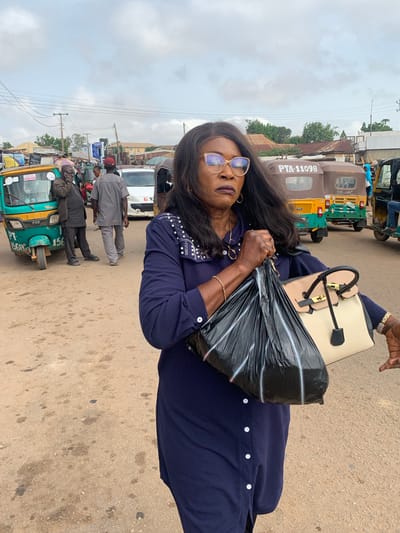 This is a full-body, eye-level, outdoor shot of a woman with dark skin, black hair, and glasses walking on a dirt road. The woman is wearing a navy blue long-sleeved dress with white buttons down the front. She is carrying a black plastic bag filled with items in her right hand and a light-colored handbag with black trim and gold hardware in her left arm. The woman's head is turned slightly to her right, and she is looking forward with a serious expression. The background of the image shows a busy street scene, with several three-wheeled vehicles, commonly known as "Keke" or "Maruwa," and other vehicles. There are also people in the background, some appearing to be vendors or passengers. The environment appears to be a market or transportation hub in Gura Top, Nigeria, characterized by a dusty road, various structures, and utility poles with wires overhead. The sky is overcast, suggesting a cloudy day.