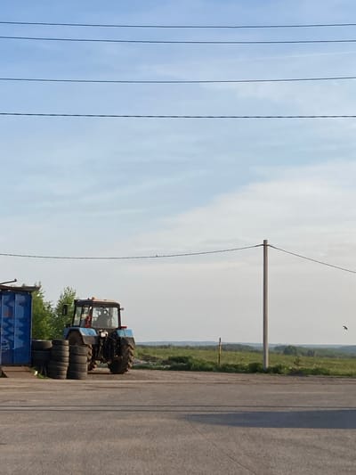 The image depicts a rural roadside scene in Verkh-Sosnova, Russia. A blue tractor with stacked tires rests near a blue trailer on a concrete road. In the background, there is a landscape of green vegetation. Overhead, power lines run parallel to the road, supported by wooden poles. A bird is visible in the sky. The sky is mostly clear with light clouds.