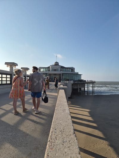 The image depicts a view of the pier in Blankenberge, Belgium, under a clear blue sky with some wispy clouds. In the foreground, a couple is walking away from the camera on the pier's walkway. The woman on the left wears a colorful, patterned dress, and the man on the right is in a grey t-shirt with a graphic design and blue shorts, carrying a black duffel bag. They are walking towards a large, circular building situated at the end of the pier, which houses a restaurant or event space with large glass windows. Other people are visible walking on the pier and near the building, as well as on the beach and in the sea. The pier's structure, with its railings and lamp posts, is visible, and shadows from the railing cast long lines across the walkway and the sandy beach to the right. The sea is moderately choppy with waves rolling onto the shore.