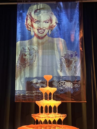The image shows a festive display featuring a large banner of Marilyn Monroe wearing large diamond earrings, set against a blue and silver background. Below the banner is a champagne tower, with several tiers of glasses filled with a yellowish liquid, illuminated from below. The banner contains text which is partially obscured by the champagne tower, but the visible words include "Marilyn Monroe with Diamond", "Herbert Dorfman", and "Corbis". The scene appears to be set up for an event, possibly a celebration or a party. There are black curtains visible on the right side of the image, suggesting an indoor setting. The text on the banner indicates this is in Wattens, Austria, likely at the Swarovski Kristallwelten, known for its crystal-themed attractions and displays.