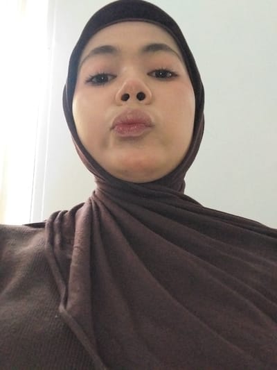 A selfie of a young woman wearing a dark brown hijab and a matching top, looking at the camera with her lips pursed. Her eyes are open, and she is wearing subtle makeup. The background is a plain, light-colored wall. The lighting suggests an indoor setting, possibly a home. The image appears to be taken from a low angle, looking up at the woman. The location in Watampone, Indonesia, is not discernible from the visual information presented.