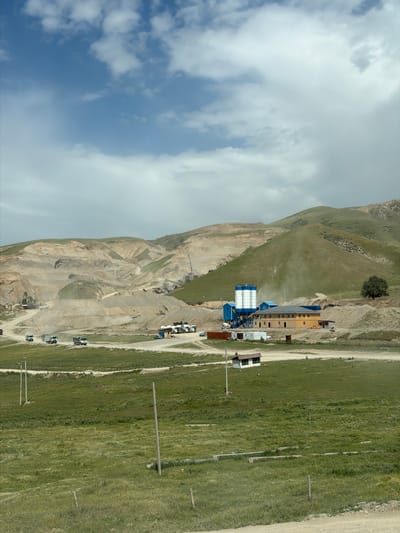 This image captures a vast, rugged landscape in Баш-Кара-Суу, Kyrgyzstan, dominated by mountainous terrain and industrial activity under a partially cloudy sky.
In the foreground and midground, expansive green grassy fields stretch out, crisscrossed by dirt roads or tracks. Several utility poles with power lines are visible, extending across these fields.
Towards the midground and background, a series of hills and mountains rise. Some slopes are covered in sparse green vegetation, while others display barren, rocky formations, suggesting a mix of soil and exposed rock. At the base of these hills, an industrial complex is situated. This complex features several buildings, including a prominent structure with large blue and white silos, indicative of a concrete plant, a quarrying operation, or a similar industrial facility. Other smaller, rectangular buildings, possibly offices or storage, are also present.
Numerous trucks, appearing to be heavy-duty or construction vehicles, are parked or moving on the dirt roads and near the industrial buildings, suggesting active operations or ongoing work related to the facility. The sky above is bright with scattered white cumulus clouds and visible patches of blue.
The scene depicts daytime, likely late morning or early afternoon. The overall impression is one of human industrial development integrated into a dramatic natural mountainous environment. No discernible text is visible on any objects or structures.