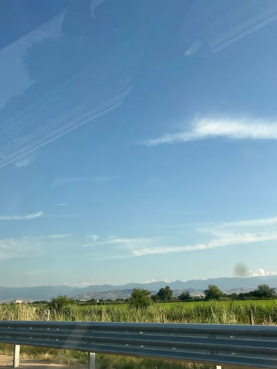 The image, taken in Dobrovnitsa, Bulgaria, depicts a scenic view from a vehicle, likely a car, as suggested by the reflection of what appears to be a car interior on the left side of the frame. The scene is dominated by a bright blue sky with scattered wispy white clouds. In the distance, a serene landscape unfolds, characterized by rolling hills covered in lush green vegetation and trees. A hazy, mountainous horizon stretches across the background, suggesting a clear, sunny day.

The foreground features a metal guardrail running along the right edge of the image, indicating the presence of a road. Beyond the guardrail, a field of tall green grass and some dry, golden grasses are visible. The overall impression is one of a peaceful, rural setting, possibly a roadside view during a journey. The time of day appears to be daytime, given the brightness of the sky and the illumination of the landscape. There is no discernible activity or event taking place; the image captures a static moment of the environment. No text is visible in the image.