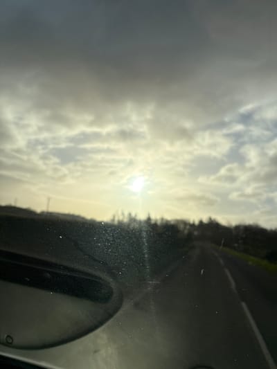 The image, taken from inside a vehicle, shows a view through a windshield. The exterior scene includes a cloudy sky with a visible sun, a roadside edge with trees, and a paved road. The interior of the vehicle is visible in the lower right corner. There is no verifiable evidence to place the location in Baschurch, United Kingdom. There are no people visible. The actions are passive: the camera is stationary.