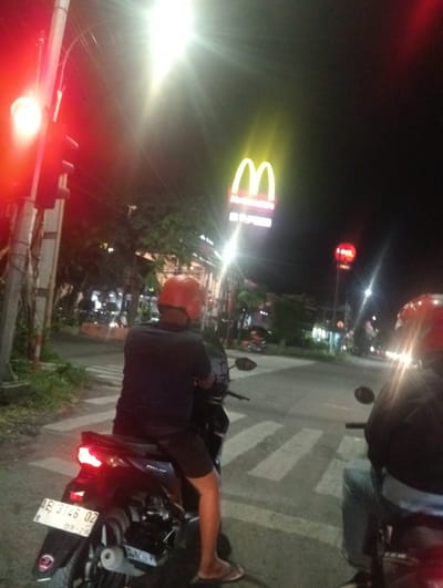 The media file captures a nighttime scene in Surabaya, East Java, featuring two individuals on motorcycles at a traffic light. The lighting suggests it is dusk or nighttime, with bright streetlights and the illuminated golden arches of a McDonald's sign prominently visible in the background.

The primary subject in the foreground is a person wearing a red helmet and a dark t-shirt, seated on a black motorcycle. This person is facing away from the camera, looking towards the traffic light which is displaying a red signal. The motorcycle has a visible license plate: "AE 3146 OZ" and "09-28".

To the right of the frame, another person on a motorcycle is partially visible, also wearing a helmet. This individual is facing the same direction as the person in the foreground.

The setting appears to be an urban street with a pedestrian crossing. Trees and buildings line the street, some of which are illuminated, indicating the presence of businesses or residences. Another illuminated sign, circular and red, is visible further down the street, possibly another advertisement or a different traffic signal. The overall atmosphere is that of a typical evening commute or a stop at an intersection in a bustling city. The bright lights and the McDonald's signage provide clear location context.